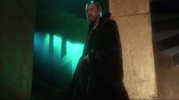 Movie still from “Excalibur” (1981), directed by John Boorman – A man in a black jacket standing next to a wall; Medium shot, Over the shoulder angle