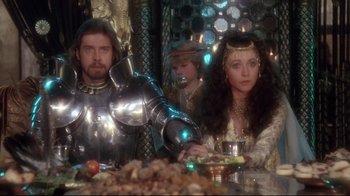 Movie still from “Excalibur” (1981), directed by John Boorman – A man and a woman sitting at a table with a child; Medium shot, Over the shoulder angle