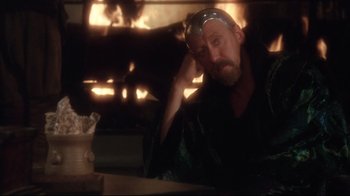 Movie still from “Excalibur” (1981), directed by John Boorman – A man sitting in front of a fire place; Medium shot, Over the shoulder angle