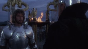 Movie still from “Excalibur” (1981), directed by John Boorman – A man in armor standing next to a fire pit; Medium shot, Over the shoulder angle