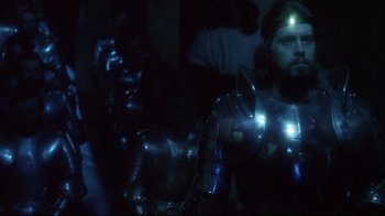 Movie still from “Excalibur” (1981), directed by John Boorman – A group of people standing next to each other in a dark room; Medium shot, Over the shoulder angle