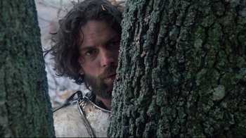 Movie still from “Excalibur” (1981), directed by John Boorman – A bearded man in a white shirt is standing next to a tree; Close Up shot, Low angle