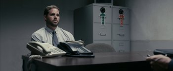 Movie still from “Fair Game” (2010), directed by Doug Liman – A man sitting at a table with a lot of office equipment; Medium shot, Low angle