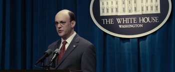 Movie still from “Fair Game” (2010), directed by Doug Liman – A man standing at a podium in front of a white house sign; Close Up shot, Over the shoulder angle