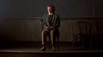 Movie still from “Fame” (1980), directed by Alan Parker – A man sitting on a chair in front of a wooden wall; Wide shot, Low angle