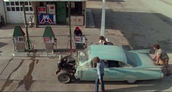 Movie still from “Fandango” (1985), directed by Kevin Reynolds – A couple of people standing next to an old car at a gas station; Wide shot, High angle