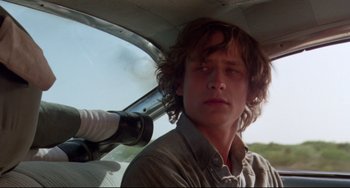 Movie still from “Fandango” (1985), directed by Kevin Reynolds – A young man with long curly hair is standing next to an airplane; Close Up shot, Low angle