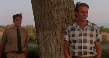 Movie still from “Fandango” (1985), directed by Kevin Reynolds – A man standing next to a tree in front of another man; Medium shot, Low angle