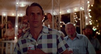 Movie still from “Fandango” (1985), directed by Kevin Reynolds – A man holding a drink in his hand while standing next to another man; Close Up shot, Over the shoulder angle