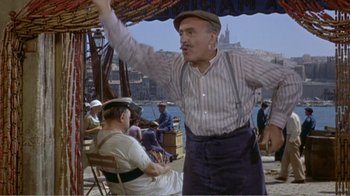 Movie still from “Fanny” (1961), directed by Joshua Logan – An older man in striped shirt and hat standing next to a man in a chair; Wide shot, Low angle