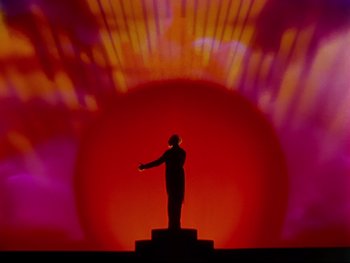 Movie still from “Fantasia” (1940), directed by Ford Beebe Jr. – A man standing on a pedestal in front of a red background; Extreme Wide shot, Low angle