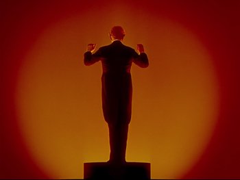 Movie still from “Fantasia” (1940), directed by Ford Beebe Jr. – A statue of a man in a black suit is lit up in a red light; Wide shot, Low angle