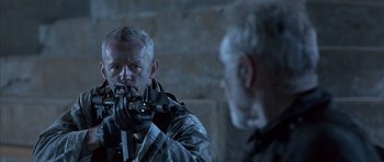 Movie still from “The Rock” (1996), directed by Michael Bay – A man holding a gun in front of another man in front of a brick wall; Medium shot, Over the shoulder angle