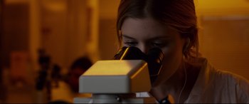 Movie still from “Fantastic Four” (2015), directed by Josh Trank – A woman looking through a microscope in a room; Close Up shot, Over the shoulder angle