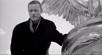 Movie still from “Faraway, So Close!” (1993), directed by Wim Wenders – A black and white photo of a man standing next to an angel statue; Medium shot, Low angle