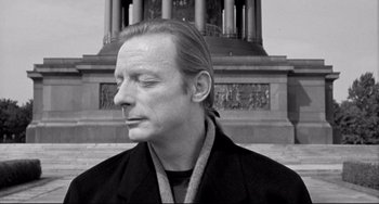 Movie still from “Faraway, So Close!” (1993), directed by Wim Wenders – Black and white photograph of a man in front of a building; Close Up shot, Low angle