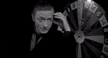 Movie still from “Faraway, So Close!” (1993), directed by Wim Wenders – A man in a black shirt and a black tie holding his hand up to his head; Close Up shot, High angle