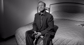 Movie still from “Faraway, So Close!” (1993), directed by Wim Wenders – A black and white photo of a man sitting on a bed; Medium shot, High angle