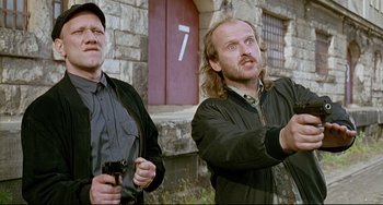 Movie still from “Faraway, So Close!” (1993), directed by Wim Wenders – Two men standing next to each other in front of a brick building; Medium shot, Low angle
