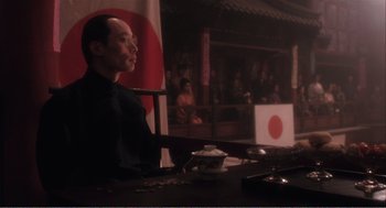 Movie still from “Farewell My Concubine” (1992), directed by Kaige Chen – A man sitting at a table in front of an asian flag; Medium shot, Over the shoulder angle