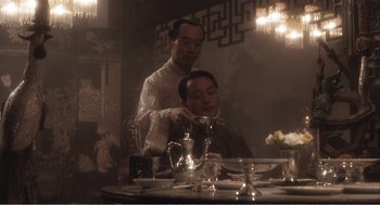 Movie still from “Farewell My Concubine” (1992), directed by Kaige Chen – A man sitting at a table with another man cutting a man's hair; Medium shot, Over the shoulder angle