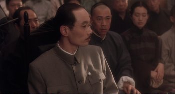 Movie still from “Farewell My Concubine” (1992), directed by Kaige Chen – A man in a suit and glasses is sitting in front of other men; Close Up shot, Over the shoulder angle