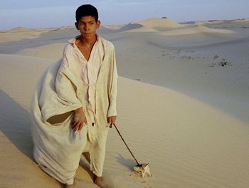 A man in a white robe and a dog on a leash in the desert