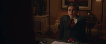 Movie still from “Fatman” (2020), directed by Ian Nelms – A man in a suit sitting at a table in front of a teddy bear; Medium shot, Over the shoulder angle