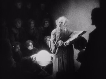 Movie still from “Faust” (1926), directed by F.W. Murnau – An old photo of a man reading a book to a group of people; Medium shot, High angle