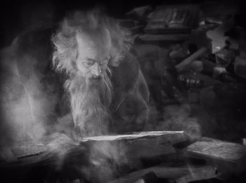 Movie still from “Faust” (1926), directed by F.W. Murnau – An old man with long hair and a long beard is looking at a piece of wood; Medium shot, High angle