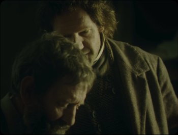 Movie still from “Faust” (2011), directed by Aleksandr Sokurov – Two men are standing next to each other in a dark room; Close Up shot, Over the shoulder angle