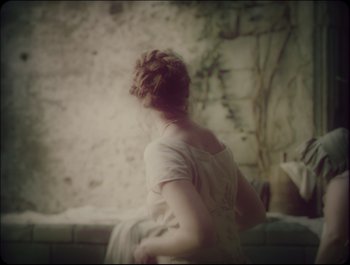Movie still from “Faust” (2011), directed by Aleksandr Sokurov – A woman in a white dress standing in front of a stone wall; Medium shot, Over the shoulder angle