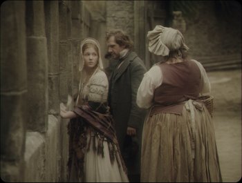 Movie still from “Faust” (2011), directed by Aleksandr Sokurov – Two women and a man in medieval garb standing next to a stone wall; Medium shot, Over the shoulder angle
