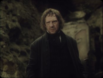 Movie still from “Faust” (2011), directed by Aleksandr Sokurov – A man in a black coat standing in front of a stone wall; Medium shot, Over the shoulder angle