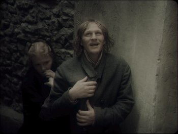 Movie still from “Faust” (2011), directed by Aleksandr Sokurov – A man standing next to a woman in a dark room; Medium shot, Over the shoulder angle