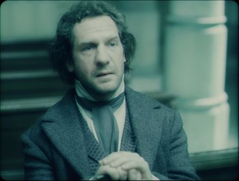 Movie still from “Faust” (2011), directed by Aleksandr Sokurov – A man in a suit and tie sitting in a room; Close Up shot, Over the shoulder angle