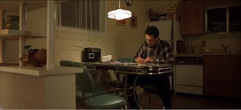Movie still from “Fear X” (2003), directed by Nicolas Winding Refn – A man sitting at a dining room table writing on a piece of paper; Wide shot, High angle