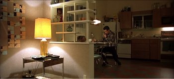 Movie still from “Fear X” (2003), directed by Nicolas Winding Refn – A man sitting at a dining room table using a computer; Wide shot, High angle
