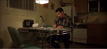 Movie still from “Fear X” (2003), directed by Nicolas Winding Refn – A man sitting at a table writing on a piece of paper; Wide shot, High angle