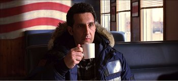 Movie still from “Fear X” (2003), directed by Nicolas Winding Refn – A man holding a coffee mug while sitting in front of an american flag; Medium shot, Over the shoulder angle