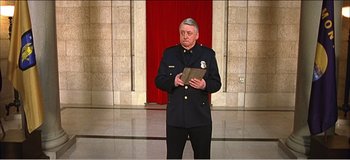 Movie still from “Fear X” (2003), directed by Nicolas Winding Refn – A man in a police uniform holding a book in front of a red curtain; Medium shot, Low angle