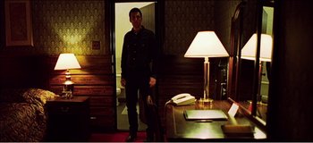 Movie still from “Fear X” (2003), directed by Nicolas Winding Refn – A man standing in front of a desk in a room; Wide shot, Low angle