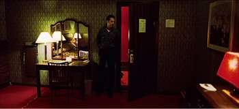 Movie still from “Fear X” (2003), directed by Nicolas Winding Refn – A man standing in front of an open door in a room; Wide shot, High angle