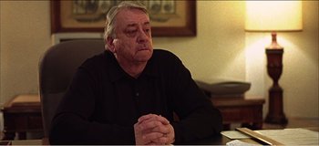 Movie still from “Fear X” (2003), directed by Nicolas Winding Refn – An older man sitting at a table with his hands clasped together; Close Up shot, Over the shoulder angle