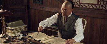 Movie still from “Fearless” (2006), directed by Ronny Yu – A man in a vest and tie holding a pair of chopsticks and pointing to a book; Medium shot, Low angle