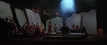 Movie still from “Fellini Satyricon” (1969), directed by Federico Fellini – A group of people sitting on a stage in front of a rock wall; Extreme Wide shot, High angle