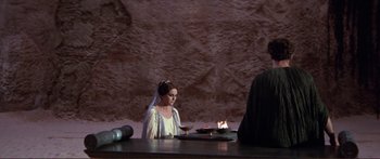 Movie still from “Fellini Satyricon” (1969), directed by Federico Fellini – A woman wearing a veil and holding a wine glass in front of a fire; Medium shot, Over the shoulder angle