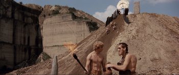 Movie still from “Fellini Satyricon” (1969), directed by Federico Fellini – Two men are standing on a hill and one is holding an axe; Wide shot, Low angle