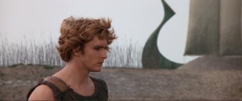 Movie still from “Fellini Satyricon” (1969), directed by Federico Fellini – A young man with blonde curly hair standing in front of a sculpture; Close Up shot, Low angle