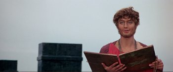 Movie still from “Fellini Satyricon” (1969), directed by Federico Fellini – A woman holding an open book while standing next to a building; Close Up shot, Low angle